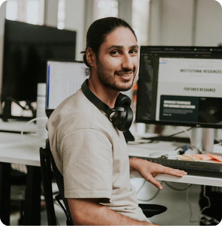 A smiling designer with headphones around his neck at his desk in front of two screens.