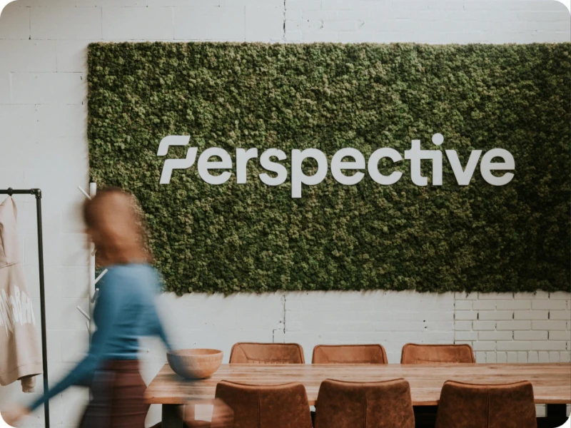 A woman walking past the Perspective logo on a green moss wall above a long wooden meeting table.