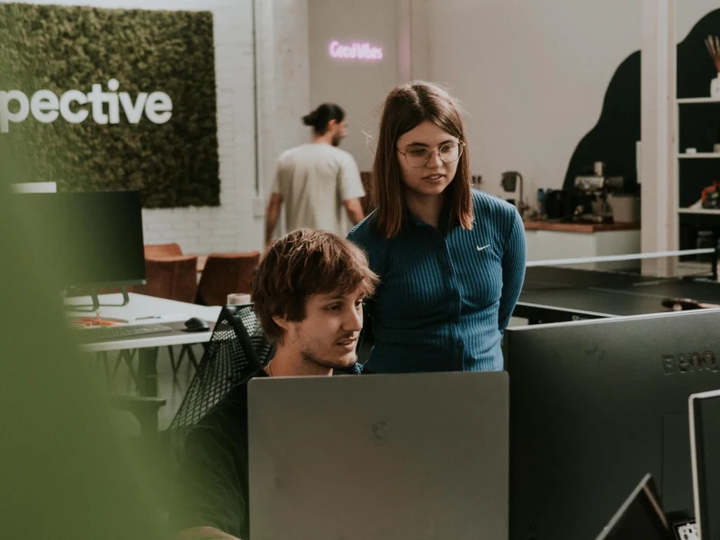 A designer standing next to a developer at his screen in the Perspective office, with the moss wall logo behind them.